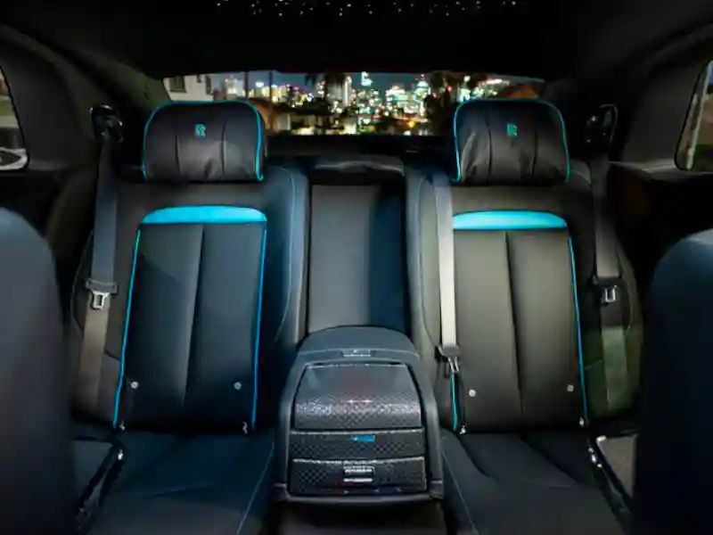 The rear passenger cabin of a Rolls-Royce Ghost Black Badge, featuring black leather seats with vibrant teal accents, embroidered RR headrests, and a glimpse of the Starlight Headliner against a night city backdrop.