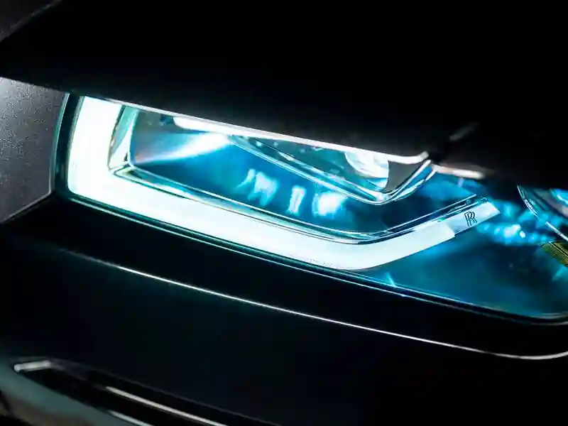 An extreme close-up of a Rolls-Royce headlight illuminating with a sharp blue glow, featuring the intricate "RR" branding etched into the glass housing.