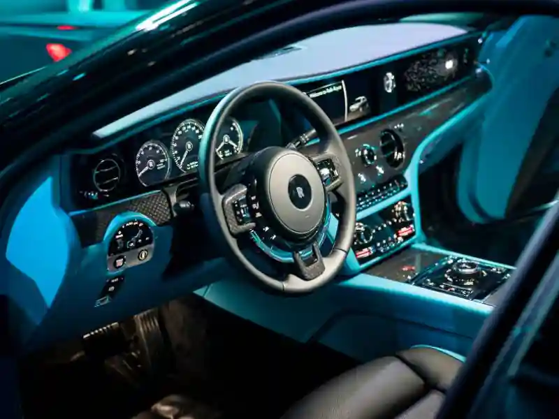 Interior cockpit of a Rolls-Royce Ghost Black Badge illuminated by vibrant blue ambient lighting, showcasing the carbon fiber dashboard trim, leather steering wheel, and digital display panels.
