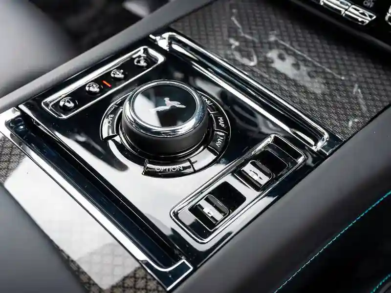 A close-up of the Rolls-Royce Ghost Black Badge center console, featuring a high-gloss black infotainment controller and Technical Fiber veneer with teal stitching on the leather armrest.