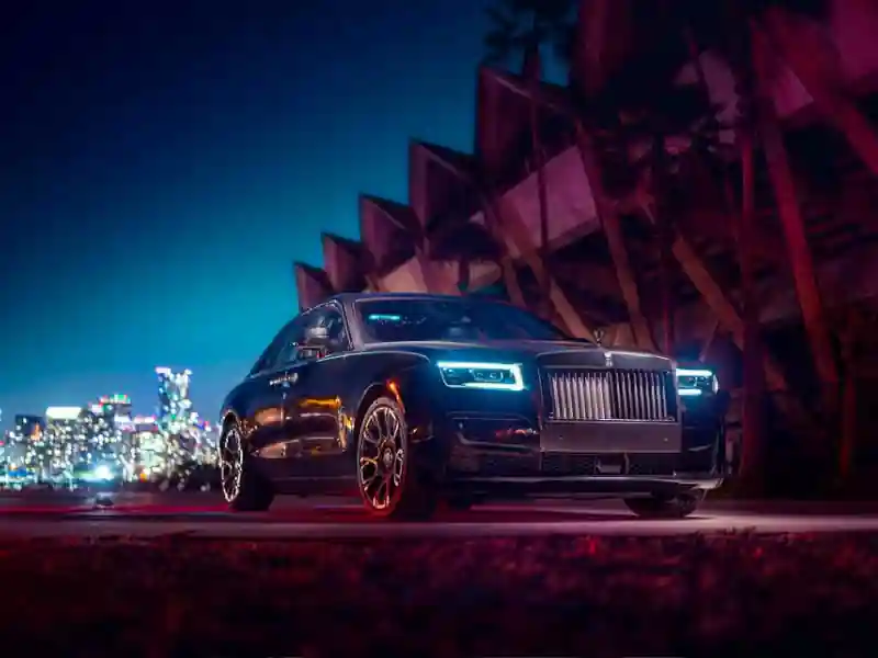 A cinematic night shot of a dark Rolls-Royce Ghost with glowing blue LED headlights, parked in front of a modern building with vibrant pink and teal neon lighting and a city skyline in the distance.