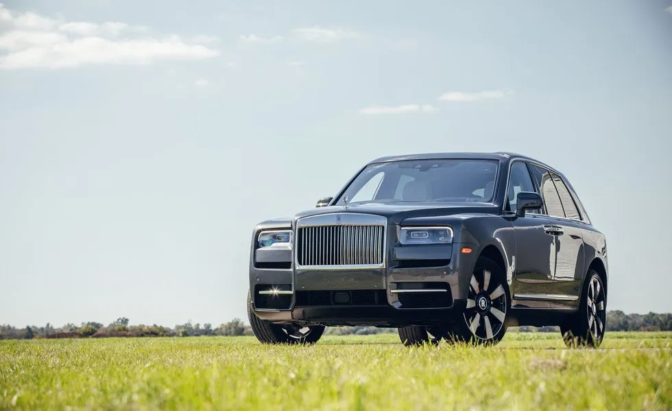A low-angle outdoor shot of a grey Rolls-Royce Cullinan parked on a lush green grass field under a bright blue sky with light clouds.