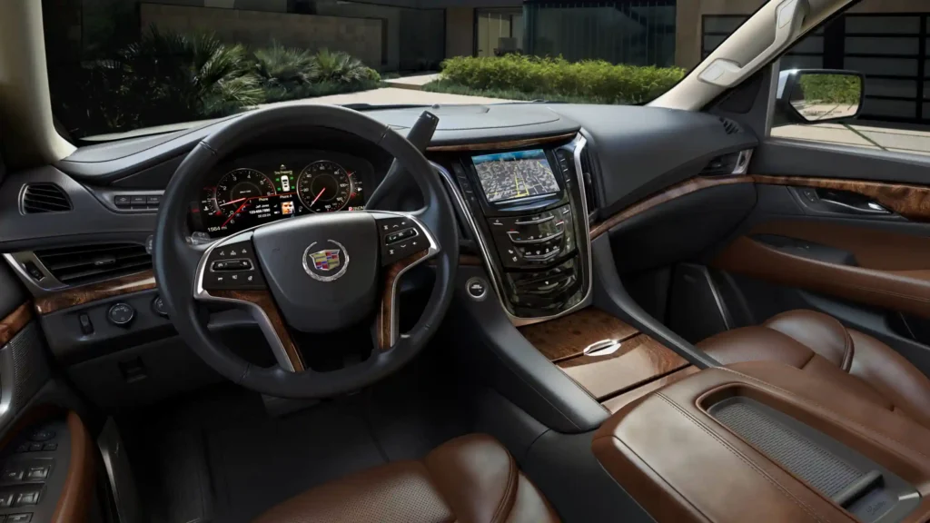 The front interior of a luxury Cadillac SUV featuring a leather-wrapped steering wheel, wood-grain trim, a digital instrument cluster, and a center console with an integrated GPS navigation system showing a city map.