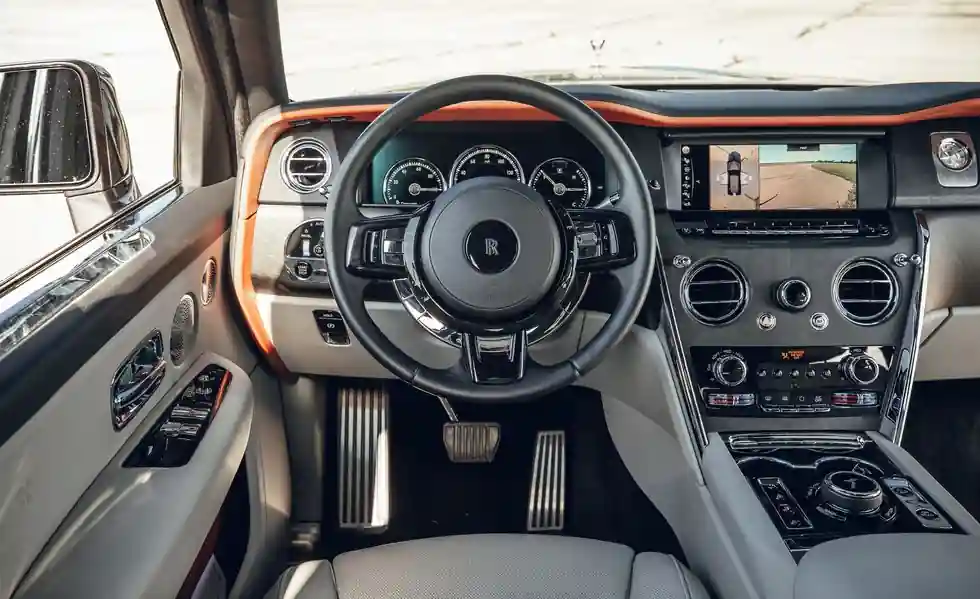 A direct view of the Rolls-Royce Cullinan driver's seat, highlighting the leather-wrapped steering wheel, digital instrument cluster, and the infotainment screen displaying a 360-degree camera view.