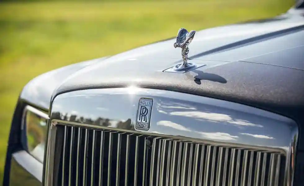 A close-up shot of the chrome Spirit of Ecstasy hood ornament and the iconic Pantheon grille of a Rolls-Royce Cullinan, with a soft-focus green background.