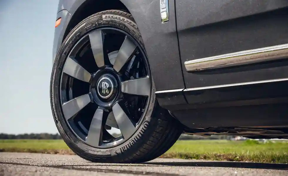 A detailed close-up of a dark grey Rolls-Royce Cullinan wheel, featuring the signature self-leveling floating "RR" hub cap and a high-performance tire on a paved road.