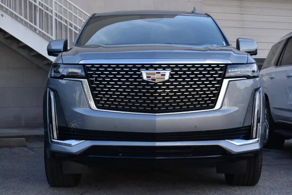 A front-view photograph of a silver Cadillac Escalade luxury SUV, highlighting its massive chrome mesh grille, iconic crest emblem, and sleek vertical LED headlights.