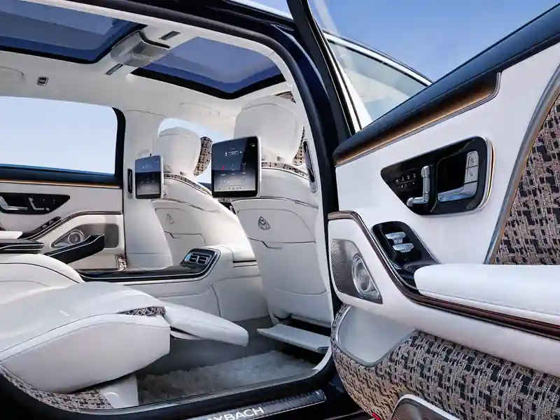 An open-door view into the rear cabin of a limited-edition Mercedes-Maybach Haute Voiture, featuring white leather reclining seats, intricate bouclé fabric door panels, dual entertainment screens, and a large panoramic sunroof.
