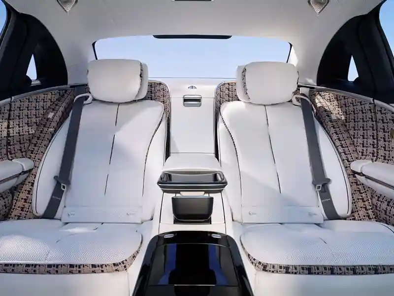 A symmetrical interior view of the rear cabin in a Mercedes-Maybach Haute Voiture, featuring white leather executive seats, patterned bouclé fabric upholstery on the doors and headrests, and a sleek black central console.