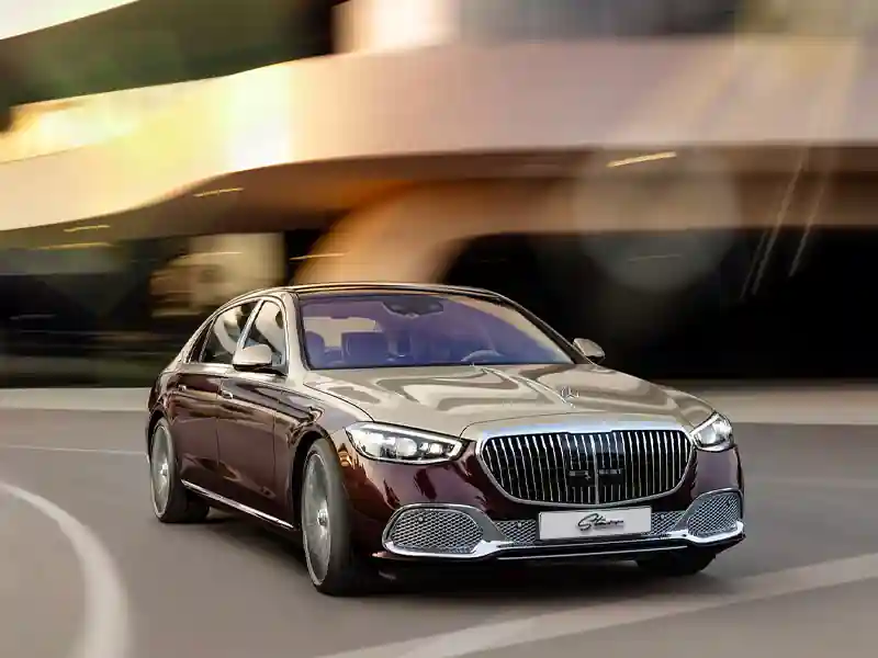 A two-tone gold and deep burgundy Mercedes-Maybach driving along a city road at sunset, with a motion-blur background of modern architecture, highlighting the large chrome vertical-slat grille and elegant LED headlights.