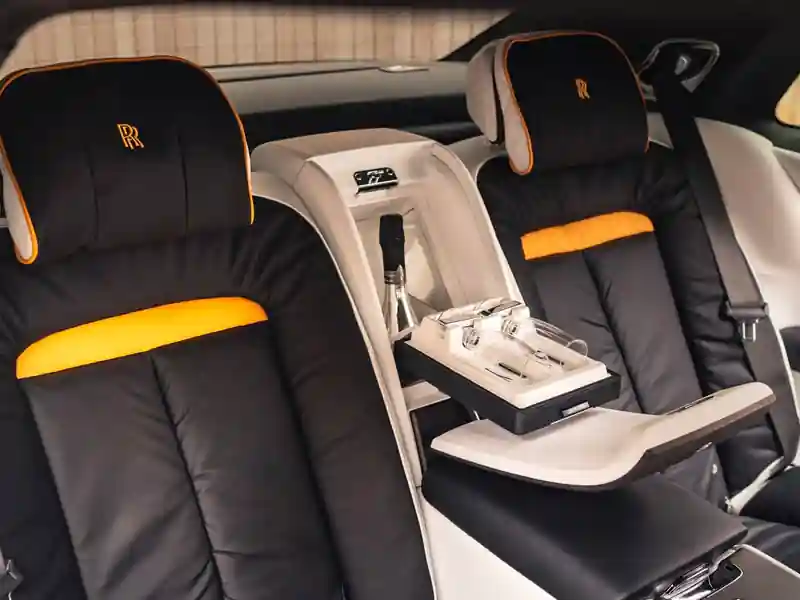 The rear seating area of a Rolls-Royce, featuring black leather seats with vibrant orange accents, embroidered RR headrests, and a center champagne cooler with crystal flutes and a bottle of bubbly.
