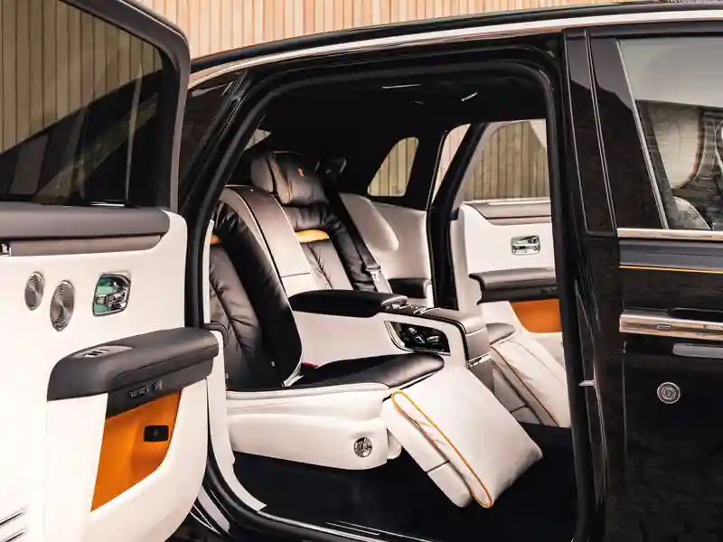 A detailed view of the luxury champagne cooler in the rear of a Rolls-Royce, featuring a bottle of champagne in a chilled compartment and two crystal glasses on a fold-down leather armrest.