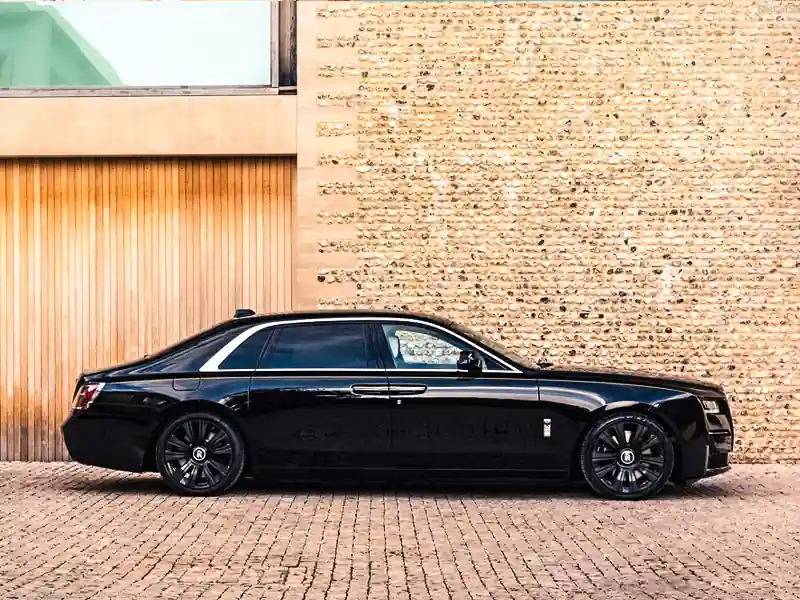 A side profile view of a black Rolls-Royce Ghost with a silver-wrapped upper body and blacked-out wheels, parked on a cobblestone courtyard against a textured stone wall.