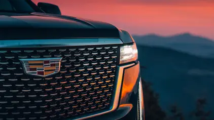 A dramatic close-up of a Cadillac Escalade's front grille and illuminated headlight at dusk, with a vibrant orange sunset and mountain range blurred in the background.