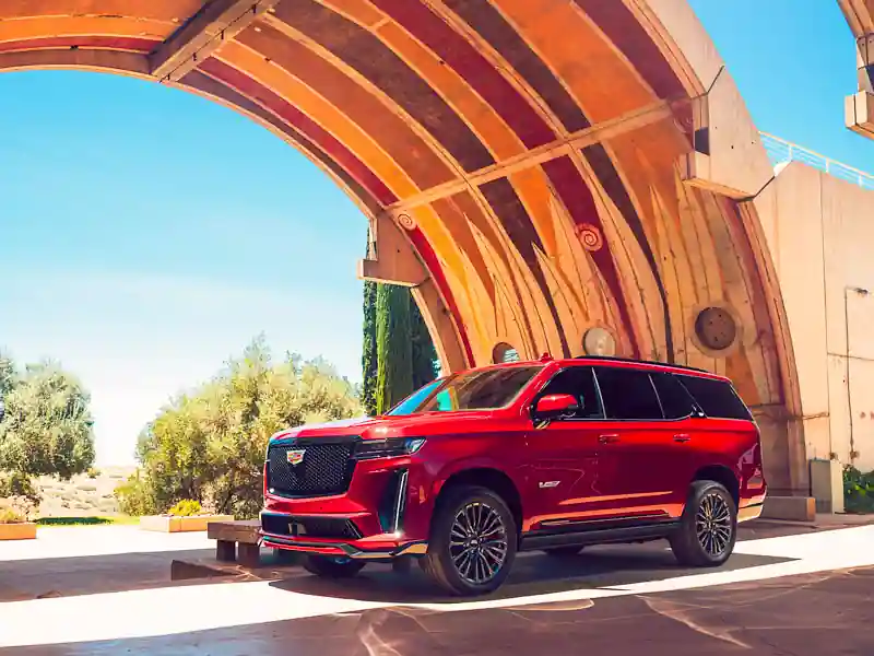 A striking cherry-red Cadillac Escalade parked under a large, modern concrete archway with artistic murals, showcasing the vehicle's bold design against a bright, sunny sky.