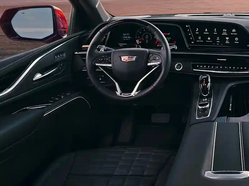 An interior view of a luxury Cadillac cockpit featuring a curved OLED digital dashboard, a leather-wrapped multi-function steering wheel, and a premium black quilted leather seat.