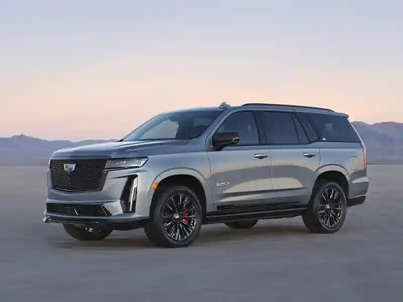 A side-profile view of a silver Cadillac Escalade-V parked on an open flat landscape with mountains in the background, featuring blacked-out rims, red brake calipers, and tinted windows.