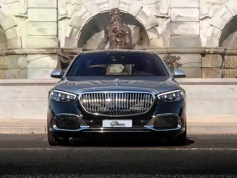 A front-view photograph of a two-tone black and silver Mercedes-Maybach parked in front of a grand classical stone fountain with a Neptune-style statue, highlighting the car's vertical chrome grille and luxury aesthetic.