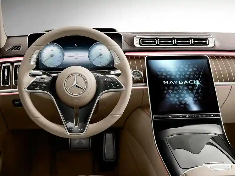 A high-tech interior view of a Mercedes-Maybach cockpit featuring a cream leather-wrapped steering wheel, a digital instrument cluster, and a large central touchscreen displaying the Maybach logo against pinstriped wood trim and ambient pink lighting.