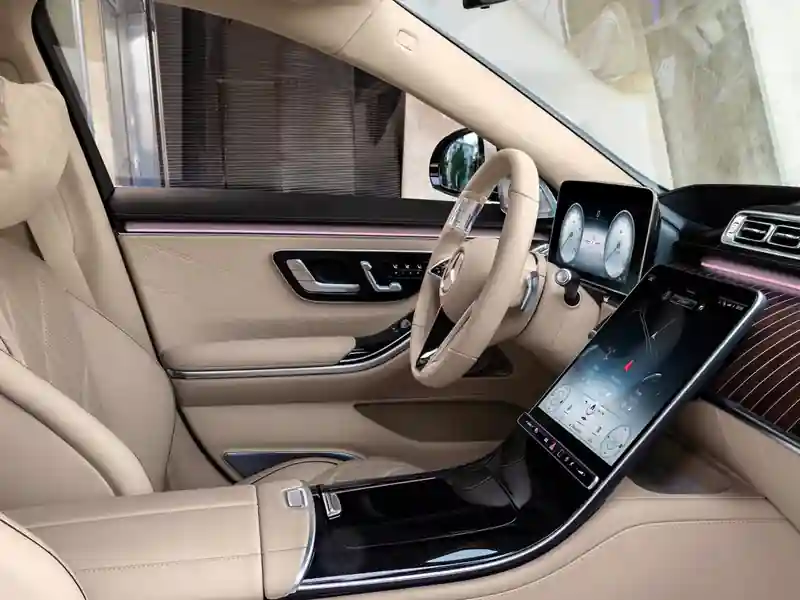 Interior side view of a Mercedes-Maybach cabin featuring diamond-quilted cream leather seats, a matching leather-wrapped steering wheel, pinstriped wood door panels, and a large central infotainment screen.
