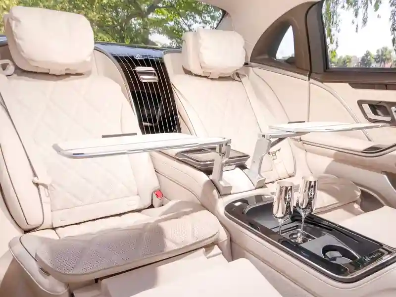 The rear cabin of a Mercedes-Maybach featuring cream diamond-stitched leather reclining seats, deployed fold-out executive work tables, and two silver champagne flutes in the center console.