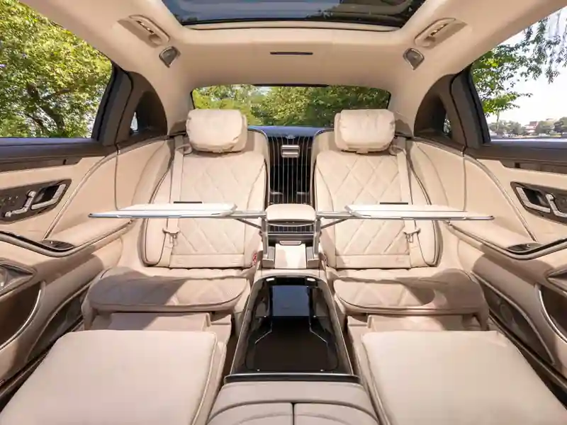 A wide-angle interior view of a Mercedes-Maybach rear cabin with both fold-out executive tables deployed, featuring cream diamond-quilted leather reclining seats and a panoramic sunroof looking out at green trees.