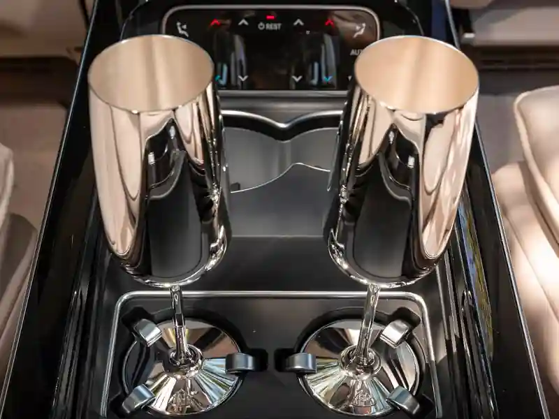 A close-up of two polished silver champagne flutes securely held in a specialized Mercedes-Maybach center console, featuring the embossed Maybach logo on the base and climate control buttons in the background.