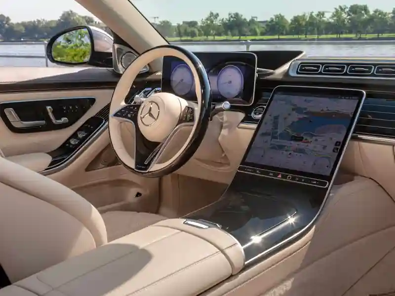 The front cockpit of a Mercedes-Maybach featuring a cream leather steering wheel and a large central touchscreen displaying a detailed GPS map, with a scenic waterfront view visible through the passenger window.
