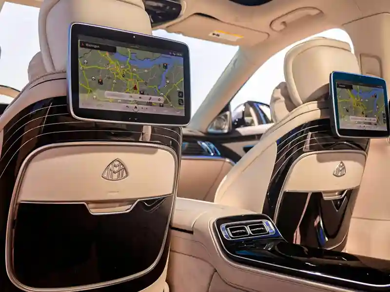 A view from the back seat of a Mercedes-Maybach featuring dual high-definition entertainment screens attached to the front headrests, displaying real-time GPS navigation maps against a cream leather and gloss-black wood interior.