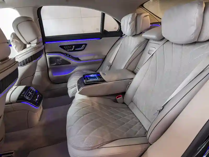 An interior view of a Mercedes-Benz S-Class rear cabin featuring light gray diamond-quilted leather seats, plush headrests, and vibrant blue ambient LED lighting along the doors and center console.