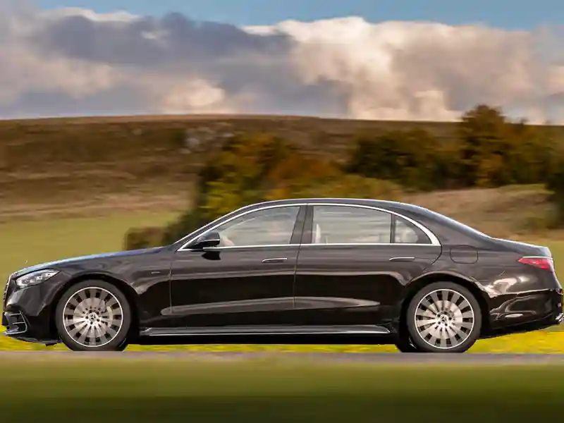 A wide-angle view of the Mercedes-Benz S-Class cabin featuring white diamond-stitched leather seats, dual rear entertainment screens, blue ambient lighting, and a panoramic sunroof.