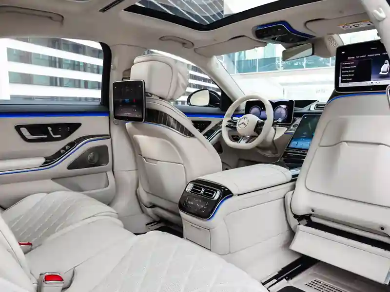 The spacious rear interior of a Mercedes-Benz S-Class featuring white diamond-quilted leather seats, dual high-definition entertainment screens, blue LED ambient lighting, and a panoramic glass roof.
