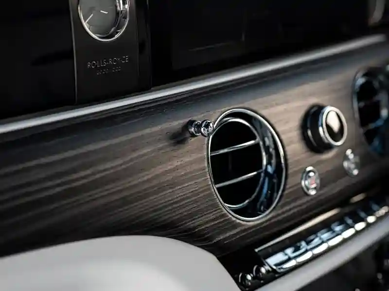 A detailed close-up of the Rolls-Royce Ghost dashboard, showcasing the texture of the open-pore wood veneer, a chrome air vent with "organ stop" control, and the signature analog clock.
