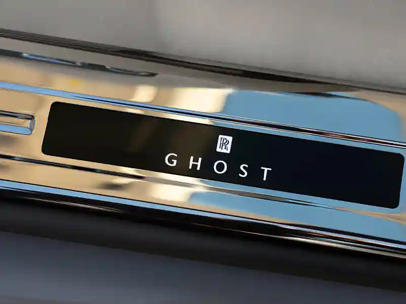 A close-up shot of the polished chrome door sill on a Rolls-Royce Ghost, featuring the illuminated "GHOST" branding and the iconic RR monogram.