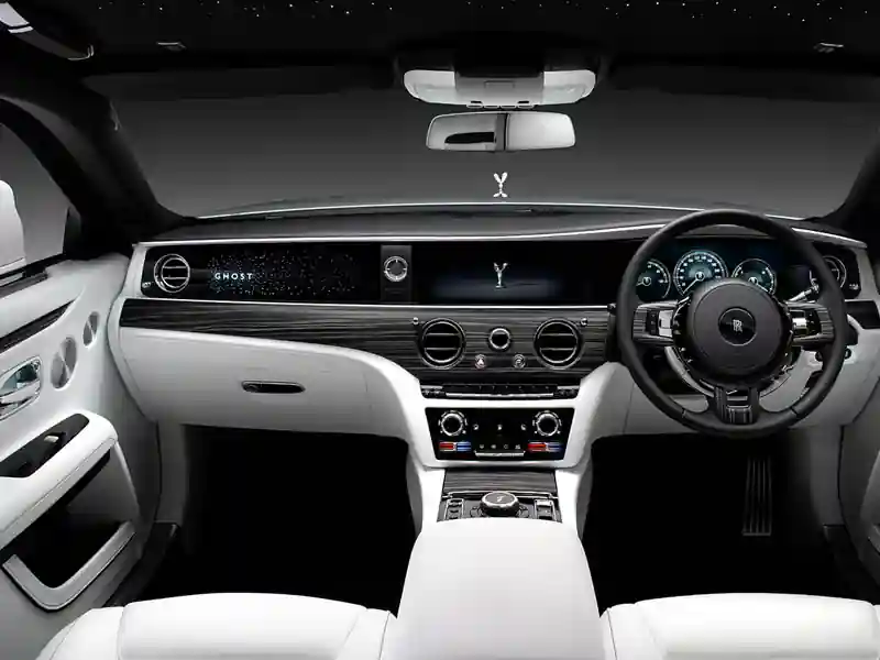 A full dashboard view of a Rolls-Royce Ghost featuring the illuminated "GHOST" passenger fascia, starlight headliner, white leather upholstery, and open-pore wood trim.