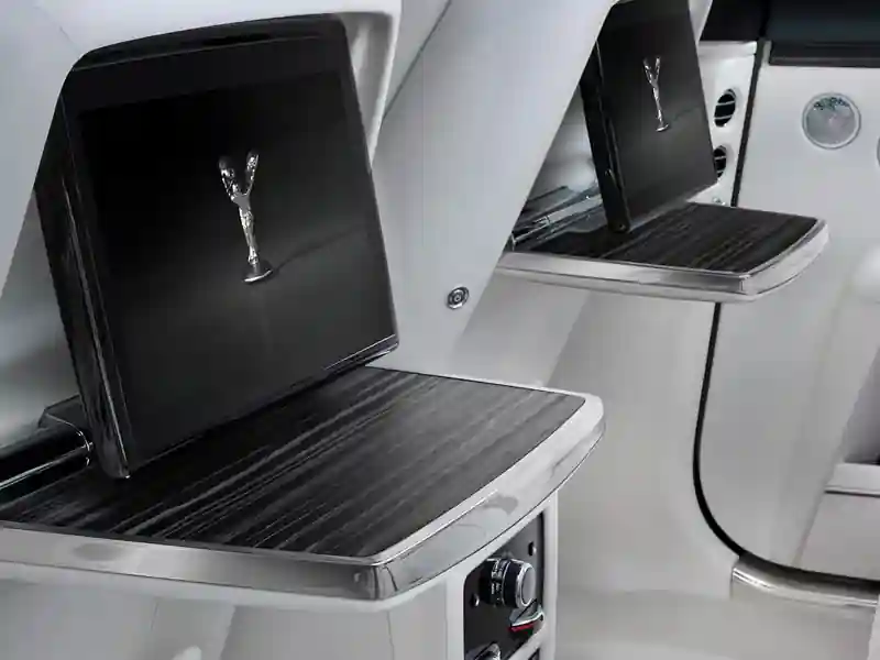 The rear passenger compartment of a Rolls-Royce Ghost, featuring dual fold-down wooden picnic tables and integrated entertainment screens displaying the Spirit of Ecstasy emblem against a white leather interior.