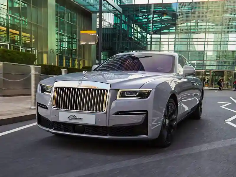 Front view of a dark Rolls-Royce Black Badge at night, highlighting the glowing white LED headlights and the imposing dark chrome vertical grille against a moody architectural backdrop.