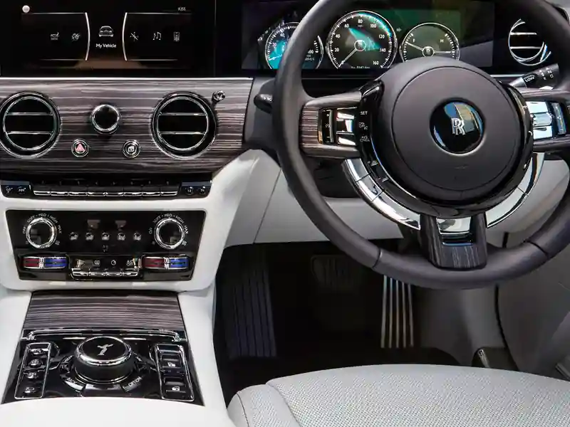 A high-resolution view of the Rolls-Royce Ghost dashboard, featuring a white leather interior, horizontal wood grain veneer, digital dials, and the rotary infotainment controller in the center console.