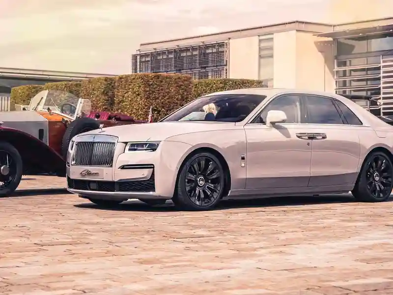 A striking blue Rolls-Royce Ghost with a silver upper body parked on a gravel driveway in front of a modern, multi-story luxury villa at dusk.