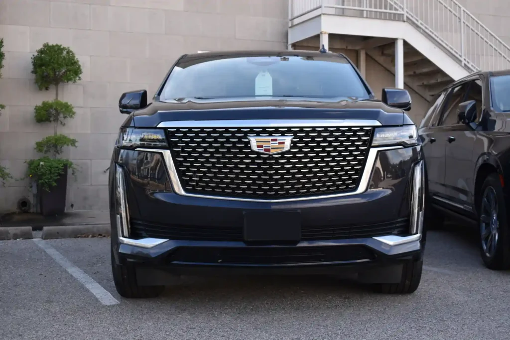 A front-view photograph of a silver Cadillac Escalade luxury SUV, highlighting its massive chrome mesh grille, iconic crest emblem, and sleek vertical LED headlights.