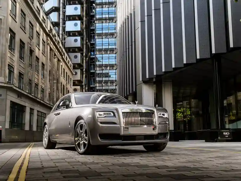 A silver Rolls-Royce Ghost parked on a cobblestone city street, positioned between a classic stone building and a modern glass-and-steel skyscraper.