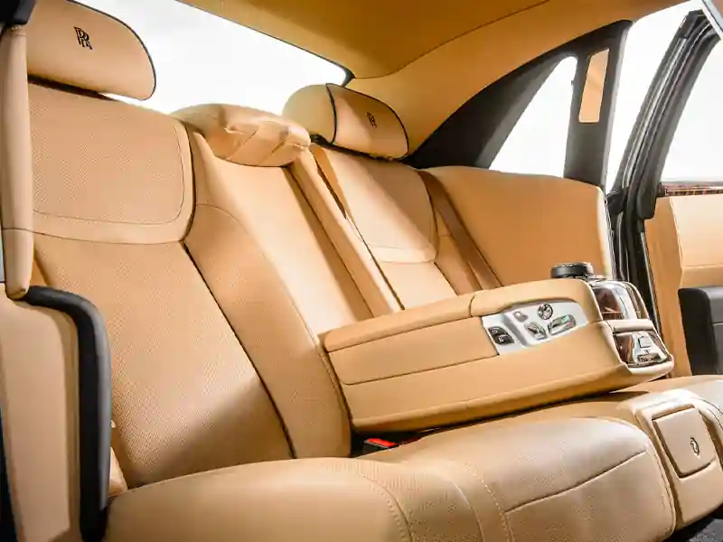 Interior view of the rear passenger cabin in a Rolls-Royce, featuring premium tan perforated leather seats, embroidered RR headrests, and a center console with seat adjustment controls.