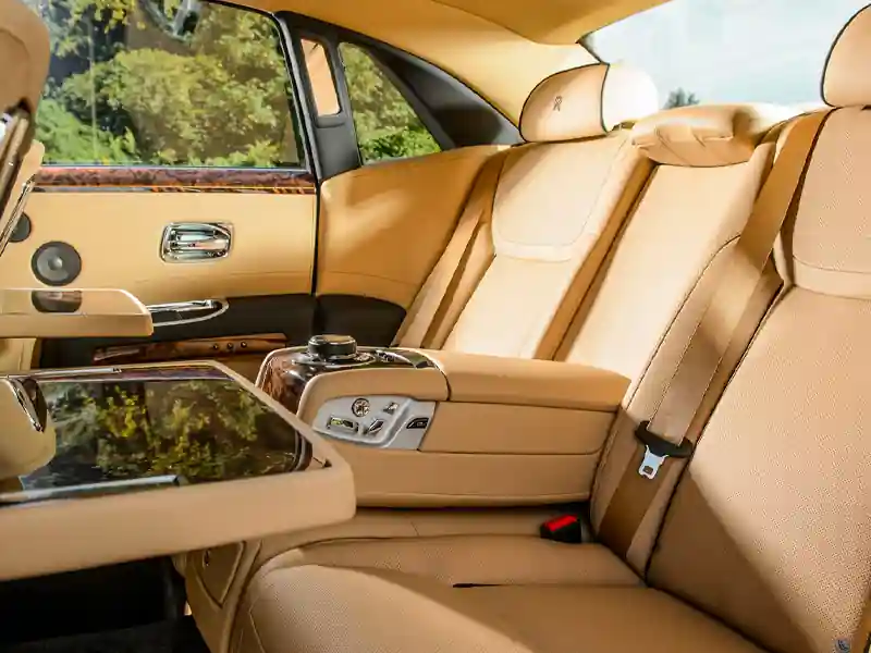 The rear luxury suite of a Rolls-Royce, showcasing fold-down wooden picnic tables, tan leather lounge seats, and high-quality speaker integration in the door panels.