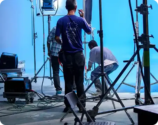 A film production crew working on a professional studio set with a large blue screen background, featuring lighting rigs, C-stands, power generators, and various technical equipment for high-end media production.