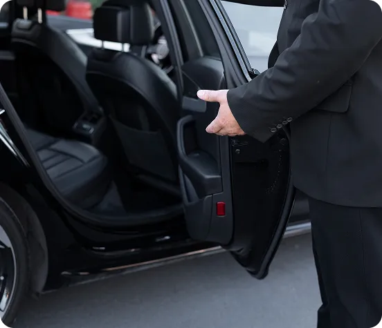 A close-up shot of a chauffeur in a formal black suit opening the rear door of a luxury black sedan, gesturing for a passenger to enter the high-end leather interior.