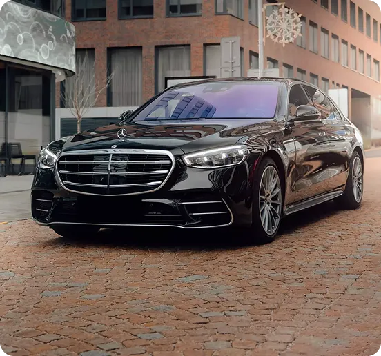A sleek black Mercedes-Benz S-Class sedan parked on a cobblestone city street in front of modern architecture, featuring a distinctive chrome grille and multi-spoke alloy wheels.