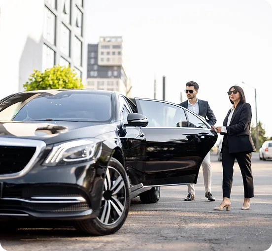 A professional female chauffeur in a black suit holding the rear door open for a male executive boarding a luxury black Mercedes-Benz S-Class in a bright urban setting.