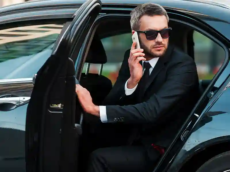 An elegant man dressed in a black suit, coming out of the Mercedes Chauffeur Service, with his mobile talking