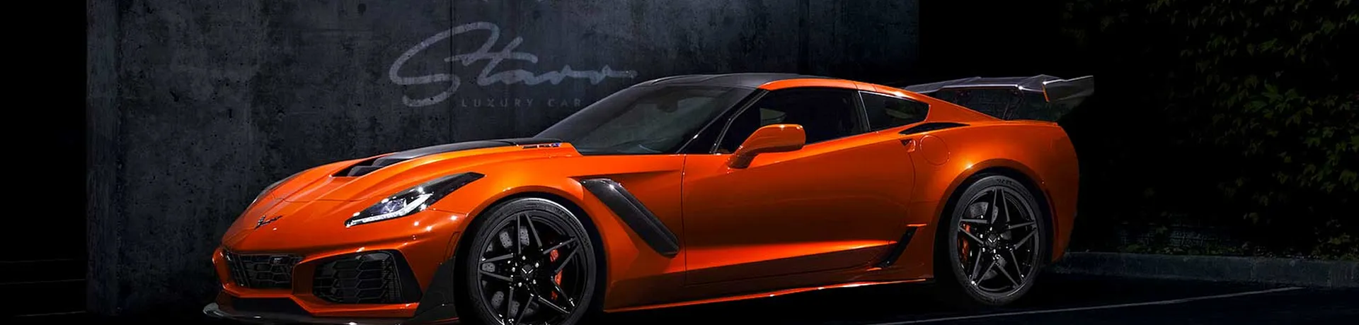 A high-performance orange Chevrolet Corvette ZR1 with a massive carbon fiber rear wing and black racing wheels, parked in front of a dark concrete wall featuring the Starr Luxury logo.