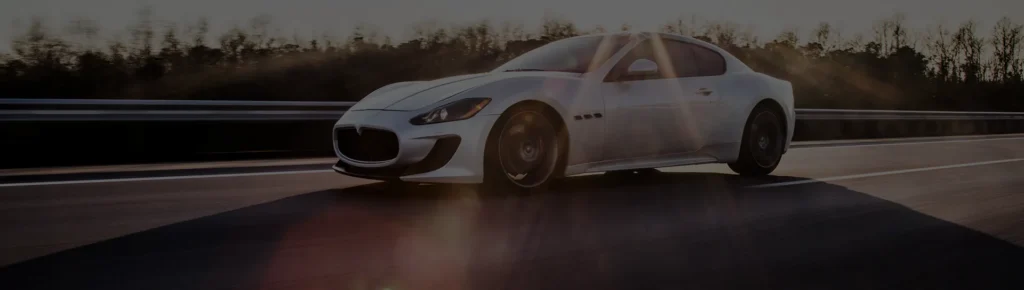 A white Maserati GranTurismo sports car speeding along a highway at sunset, featuring dramatic sun flare, motion blur, and a dark, moody landscape background.